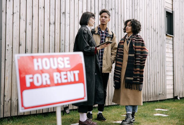 Three people talking outside behind a house for rent sign